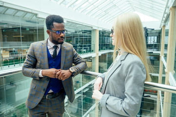 african american man in a white shirt in brown jacket posing talking to a business blonde caucasus woman in a gray suit in a modern office with glass windows shopping mall
