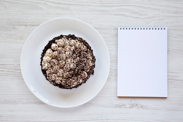 Homemade tiramisu cake on a white plate, blank notepad on a white wooden surface, top view. Copy space.