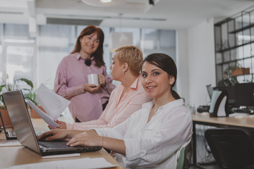 Obraz premium Group of successful businesswomen working at the office together. Lovely young businesswoman smiling to the camera, her female colleagues talking on the background. Startup, equality concept