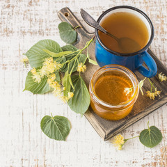 A small glass jar with honey, fresh linden flowers and a mug of hot tea on a square wooden board on a light background. Prevention and treatment of flu and colds. Selective focus.