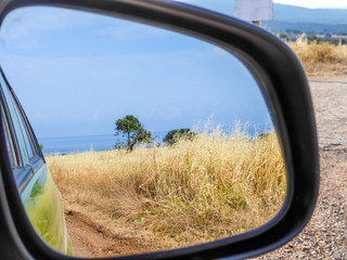 A beach view seen in the side mirror of a car. Driving through a graveled road. Ground is overgrown with a dried grass. Some trees growing next to the beach.