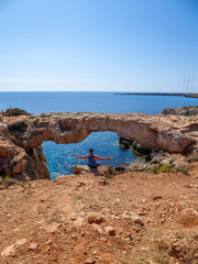 A young man sitting at the steep cliff of Cape Greco, Cyprus, in front of his is a stony arch, bridge connecting two cliffs. Barren slopes of the stony formation. Blue sea. Endless line of a horizon.