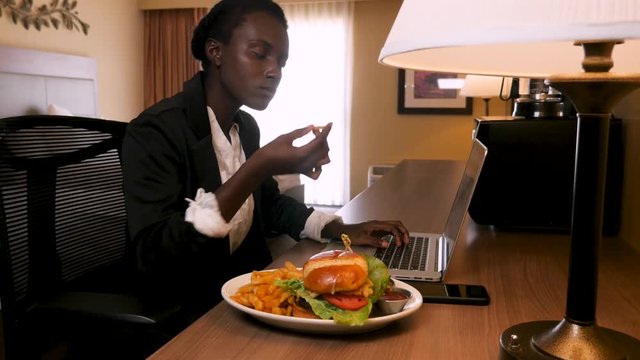 Business Woman Working And Eating In Hotel Room. Inside her hotel room, this African decent woman works on her laptop. After typing a bit she picks up a french fry next to her gourmet hamburger and ea