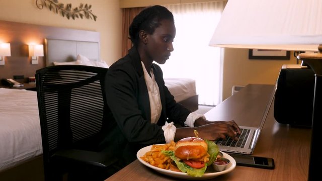 business woman working at hotel desk while eating food.  Inside her hotel room, this African decent woman works on her laptop. After typing a bit she picks up a french fry next to her gourmet hamburge
