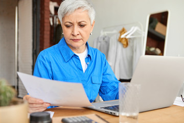 People, lifestyle, domesticity and modern technology concept. Concentrated retired woman with short gray hair holding sheet of paper, doing domestic finances at home using laptop and calculator
