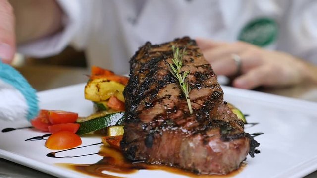 chef dabs meat juice on plate slow motion. The chef reaches around to the front of the white dish to sop up the extra juice from the steak on the plate as the camera slowly moves side to side.