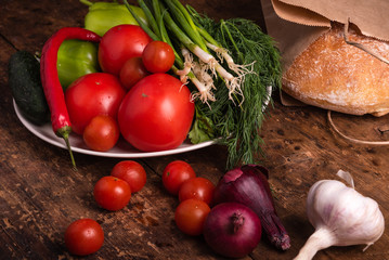 Fresh vegetables, tomatoes, cherry tomatoes, green peppers, chilli peppers, onions, cucumbers, garlic and herbs and ciabatta bread in a paper bag on a rustic wooden table - Italian rural style