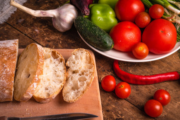 Fresh vegetables tomatoes, onions, peppers, cucumbers, garlic and herbs on and ciabatta bread cut on a rustic wooden table - italian rustic style