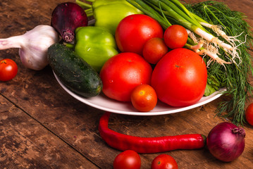Fresh vegetables, tomatoes, cherry tomatoes, green peppers, chilli peppers, onions, cucumbers, garlic and herbs in a plate on a rustic wooden table - rural style, healthy, organic food concept