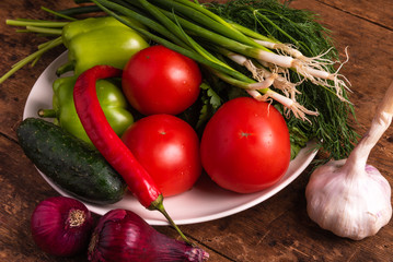 Fresh vegetables, tomatoes, cherry tomatoes, green peppers, chilli peppers, onions, cucumbers, garlic and herbs in a plate on a rustic wooden table - rural style, healthy, organic food concept