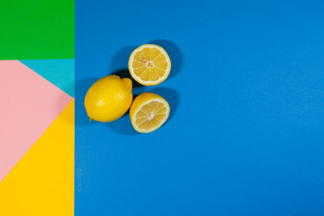 Two lemons, one whole and one cut in half, laying on a flat surface of different colors pink, yellow, blue, and green