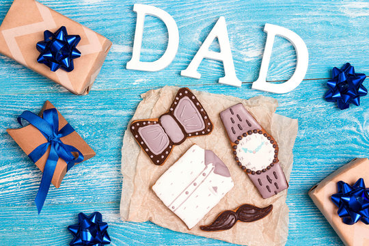 Tasty creative cookies with word dad and gift boxes on blue wooden background. Gingerbread in the shape of a shirt, watch, bow tie and mustache.