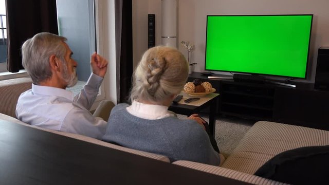 An elderly couple sits on a couch in a living room, watches TV with a green screen and talks