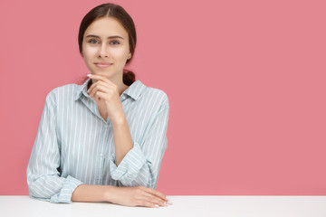 Cute woman employee in striped shirt touching chin and smiling, posing isolated against pink wall...