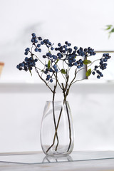 A shot of an interior design. In the middle there is a composition with several blueberry twigs in a transparent tear-shaped vase. On the blurred background there is a wall shelf with a photo frame.