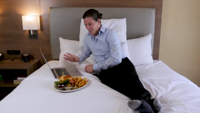 Man On Hotel Bed Speaking To Laptop With Food. Man speaks to his laptop computer as he lays on his hotel bed dressed in business attire with a man bun. He smiles as he speaks then picks up a fry from 