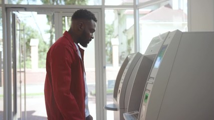 Side view of afro-american attractive man using ATM machine inserting a credit card and entering pin indoors.