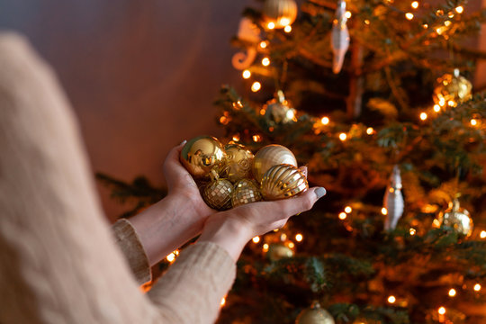 Close-up woman holds in the hands of Golden Christmas tree toys. Winter holidays in a house interior. Golden and white Christmas toys, lights garlands. Natural Danish spruce