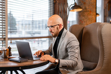 Portrait of young businessman in comfortable cafe. Man with beard in the glasses looking to the monitor laptop. Successful people. Reading text during work on netbook
