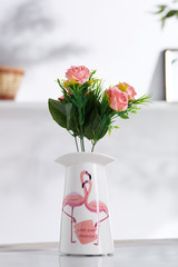 Shot of interior design. The floral composition is in middle, with bunch of pink roses in a porcelain vase with literal and bird print. On blurred background there is wall shelf with photo frame.
