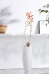 Shot of interior design. There is floral composition in middle with immortelle twigs in ceramic vase with skewed opening. On blurred background there is wall shelf with photo frame and woven basket.