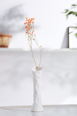 A shot of interior design. There is floral composition in middle with immortelle twigs in vase with widened opening and relief surface. On blurred background there is wall shelf with a photo frame.