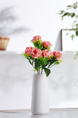Closeup shot of an interior design. There is floral composition in middle with pink roses in a snowy bisque vase with smooth round surface. On blurred background there is wall shelf with photo frame.