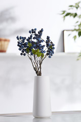 Closeup shot of interior design. There is floral composition in middle with blueberry twigs in snowy bisque vase with smooth round surface. On blurred background there is wall shelf with photo frame.