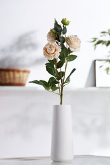 Closeup shot of an interior design. There is floral composition in middle with China roses in snowy bisque vase with smooth round surface. On blurred background there is wall shelf with photo frame.