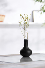 Shot of interior design. There is floral composition in the middle with immortelle twigs in a black bud vase with high neck and wide body. On blurred background there is wall shelf with photo frame.