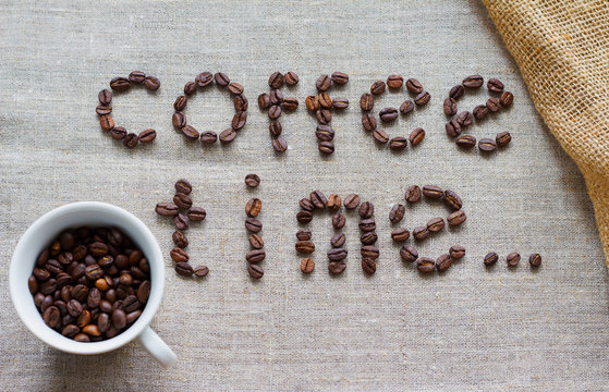 Inscription Coffee Time of a roasted coffee beans on a grey canvas, white cup of coffee beans at bottom left corner table,  sackcloth at upper right