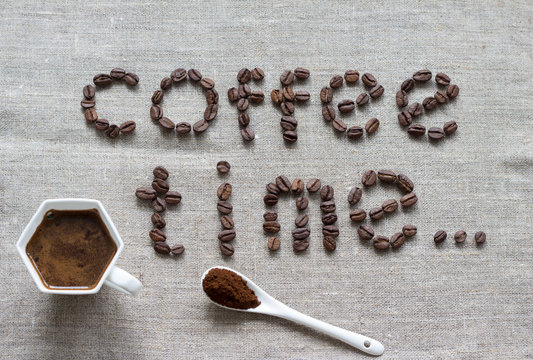 Inscription Coffee Time of a roasted coffee beans on a grey canvas, white sixangle porcelain cup of coffee and white spoon with ground coffee, table