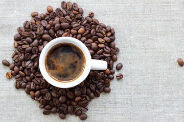 White cup of coffee within a roasted coffee beans on a grey textile background, top view, left side