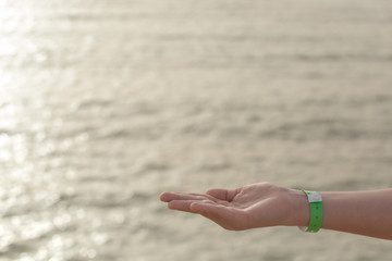Fototapeta na wymiar Closeup view of kids hand wearing green entrance wristband of hotel resort or entertainment park. Hand in gesture with empty palm isolated on sea water background. Horizontal photography of tourist.
