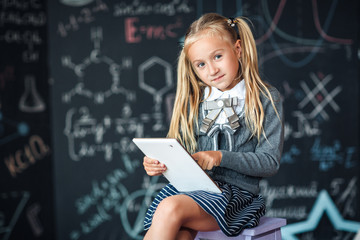 Adorable little blond schoolgirl in school uniform holding white digital tablet. Chalkboard with school formulas background. Back to school concept.