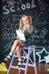 Little blond Girl in school uniform holding white tablet pc in chemistry class. Chalkboard with school formulas background.