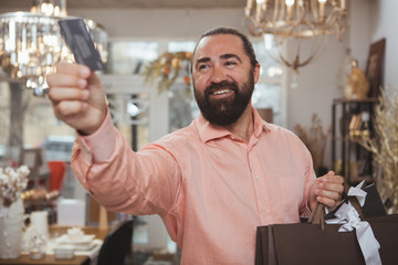 Handsome bearded mature man holding shopping bags, giving his credit card at checkout counter.Attractive male customer paying for his purchase at the store. Buying, spending money, banking concept