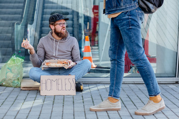 Stylish homeless beggar man in eyeglasses eating pizza while sitting on the ground with bag full of waste paper and garbage bag near the shopping mall. Concept of human indifference.
