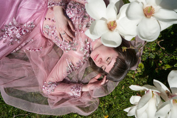 A young girl in a lush pink dress lies with her eyes closed under a magnolia tree with large white...