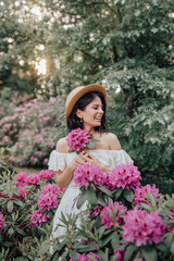 Fototapeta premium Close up portrait of a beautiful girl in a white vintage dress and straw hat standing near pink flowers