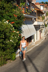 girl in shorts and a white top walks down the street in a Greek village past buildings and...
