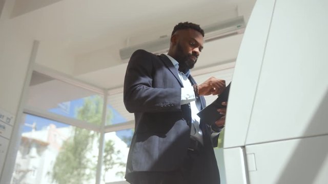Bottom view of african confident businessman withdrawing a salary from his card. Handsome man in suit inserting a credit card into an ATM machine in a bank.