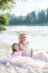Fototapeta na wymiar A cheerful child and her mother are enjoying the sunny morning near the water. Portrait of a beautiful blonde mother and her daughter in beautiful dresses on the sand with a lake in the background. 