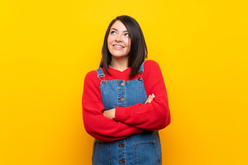 Fototapeta premium Young Mexican woman with overalls over yellow wall looking up while smiling