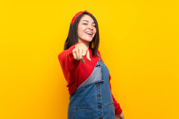 Young Mexican woman with overalls over yellow wall points finger at you with a confident expression