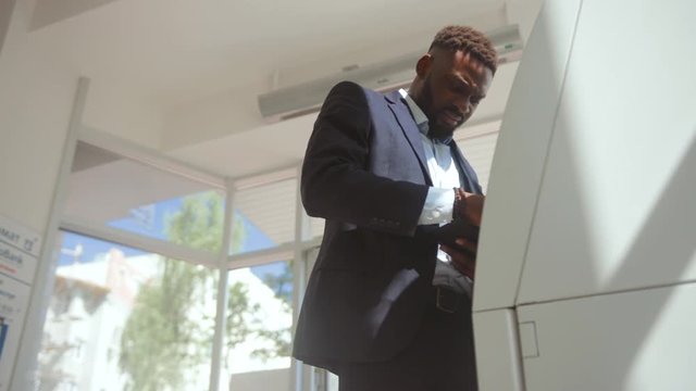 Busy african man in business suit inserting a credit card into an ATM and entering a pin code on keyboard. Side view of a serious businessman withdrawing money from a cash dispenser.