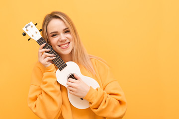 Smiling blonde in yellow clothes with ukulele in hand is isolated on an orange background, looking into the camera and rejoicing. Portrait of a positive girl with Hawaiian guitar in hand. Copy space - obrazy, fototapety, plakaty