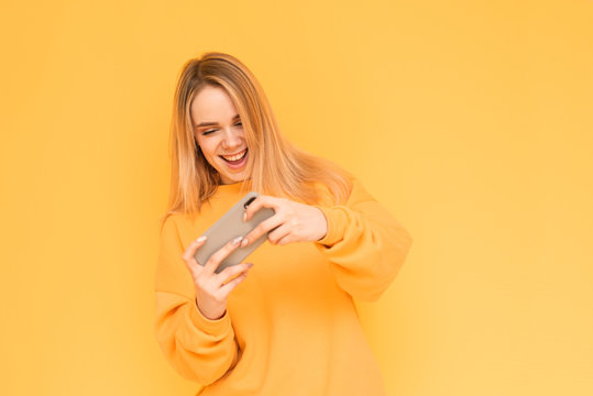 Portrait of a teenage girl in orange clothes playing mobile games on a smartphone, smiling, looking at the phone screen on a yellow background. Mobile gaming. Girl is a gamer. Isolated.
