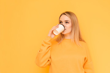 Beautiful girl in bright clothes is standing on an orange background and drinking coffee from a paper cup, looking sideways and smiling. Isolated. Copy space