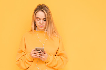 Blonde girl in orange clothing listens to music in headphones and uses a smartphone on a yellow background. Lady is using the internet on a smartphone. Isolated.